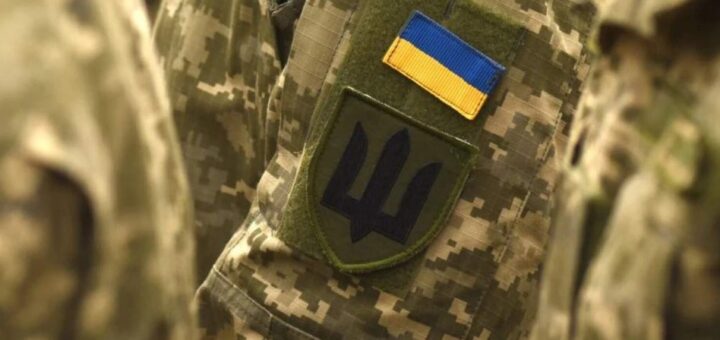Крупний план камуфльованої військової форми; на рукаві два шеврони — прапор України (синьо‑жовті смуги) та зелений щит із синім тризубом; символи Збройних Сил України.»