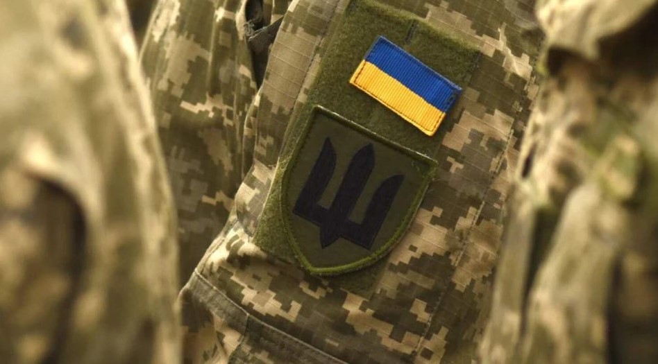 Крупний план камуфльованої військової форми; на рукаві два шеврони — прапор України (синьо‑жовті смуги) та зелений щит із синім тризубом; символи Збройних Сил України.»