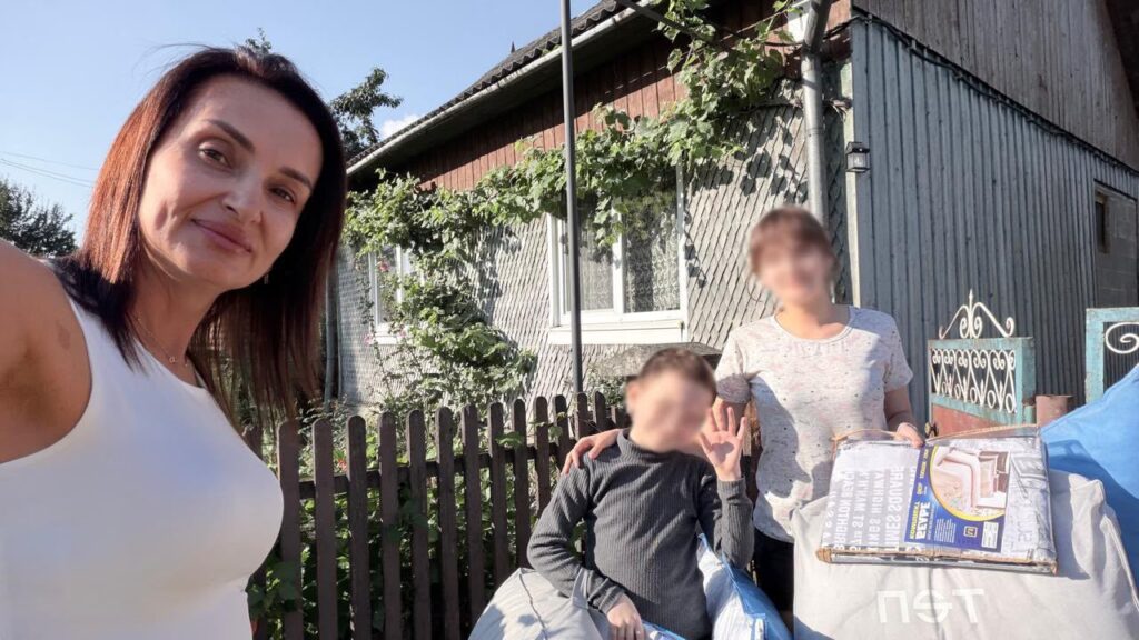 Тетяна Гасаненко та жінка з дитиною біля дерев’яного паркану та будинку з виноградною лозою. Жінка з хлопчиком тримають великі пакунки.