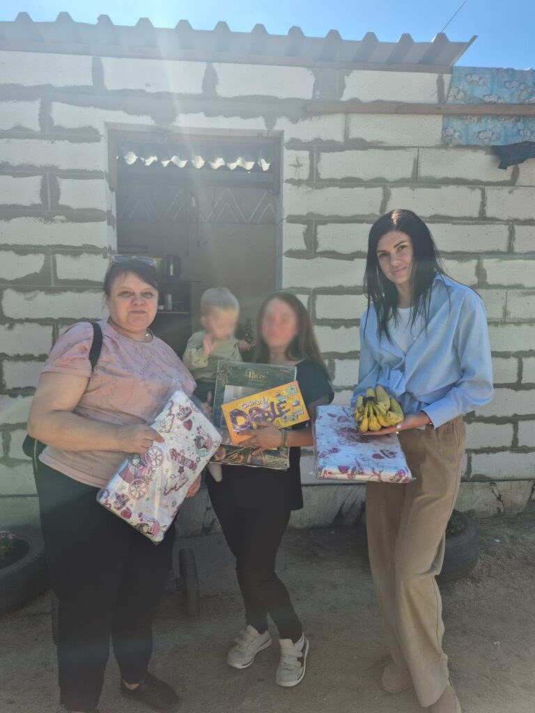 Троє жінок дорослих і дитина на руках однієї жінки біля будівлі. В руках жінки тримають коробки з подарунками та банани.