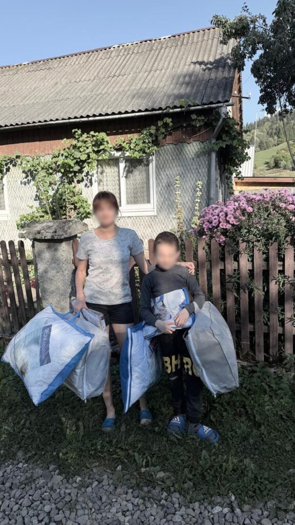 Двоє людей з великими синьо‑білими сумками стоять біля будинку з квітами та зеленими лозами.