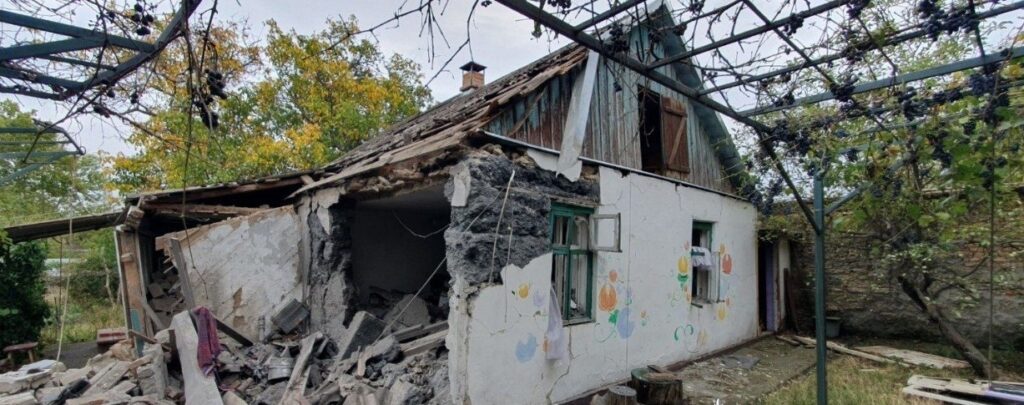 Зруйнований будинок із частково обваленим дахом і стіною, поруч виноградна арка.