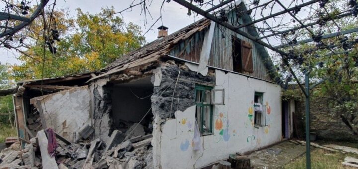 Зруйнований будинок із частково обваленим дахом і стіною, поруч виноградна арка.