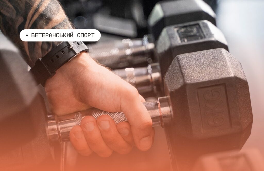 Рука з татуюваннями тримає гантелю вагою 6 кг у тренажерному залі; на задньому плані видно інші гантелі на стійці; на зображенні напис українською — “Ветеранський спорт”.