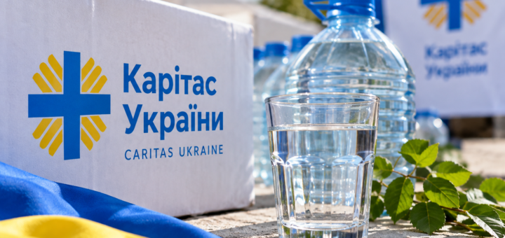 гуманітарна допомога від «Карітас України». Білий ящик із логотипом організації, поруч склянка води та гілочка зелені. На передньому плані видно український прапор, позаду — великі пляшки з водою та банер із тим самим логотипом.