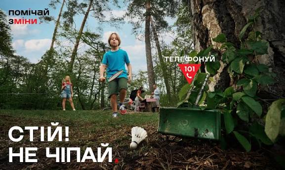 Дитина у парку помічає зелений металевий ящик біля дерева, поруч лежить волан для бадмінтону; на плакаті написано українською: «Помічай зміни», «Телефонуй 101», «Стій! Не чіпай».