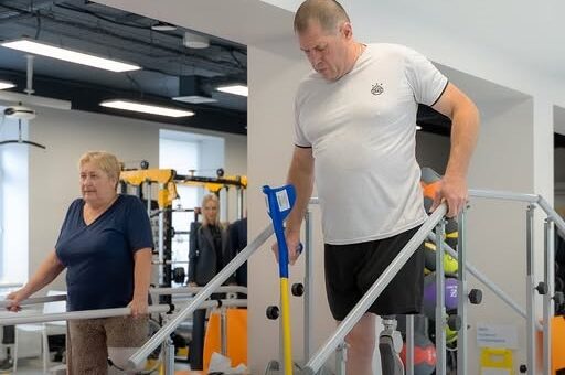 Двоє людей виконують вправи на паралельних брусах у сучасному реабілітаційному центрі. Один із них пересувається з милицею, інший тренує рівновагу.