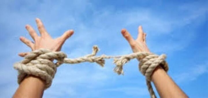 Дві руки підняті до неба, на зап’ястях розірваний канат, що символізує свободу та звільнення. На фоні — чисте блакитне небо з легкими хмарами.