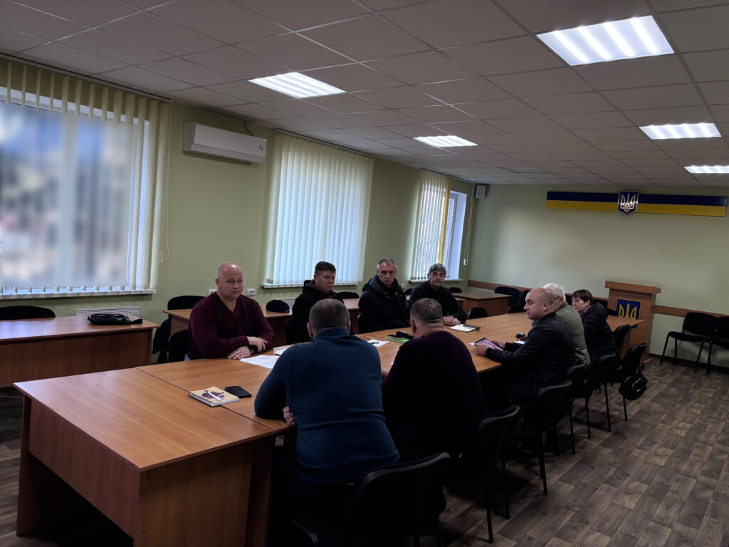 вісім людей за великим столом у світлій кімнаті з українським гербом і синьо‑жовтою смугою на стіні — офіційне засідання.