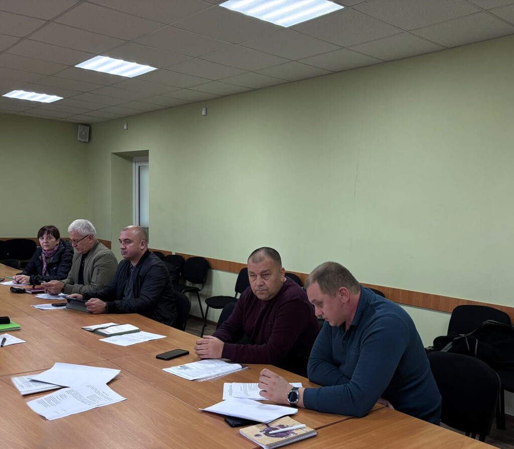 п’ятеро людей сидять за довгим дерев’яним столом у кімнаті з зеленими стінами, на столі папери й телефони — робоча зустріч.